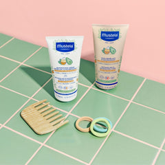 Two Mustela baby care products on a green and pink checkered surface with a wooden comb and rubber rings.