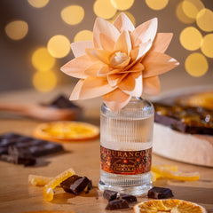 Small bottle of Scented Flower with a decorative flower on a wooden surface with dried oranges and chocolates, blurred lights in the background.