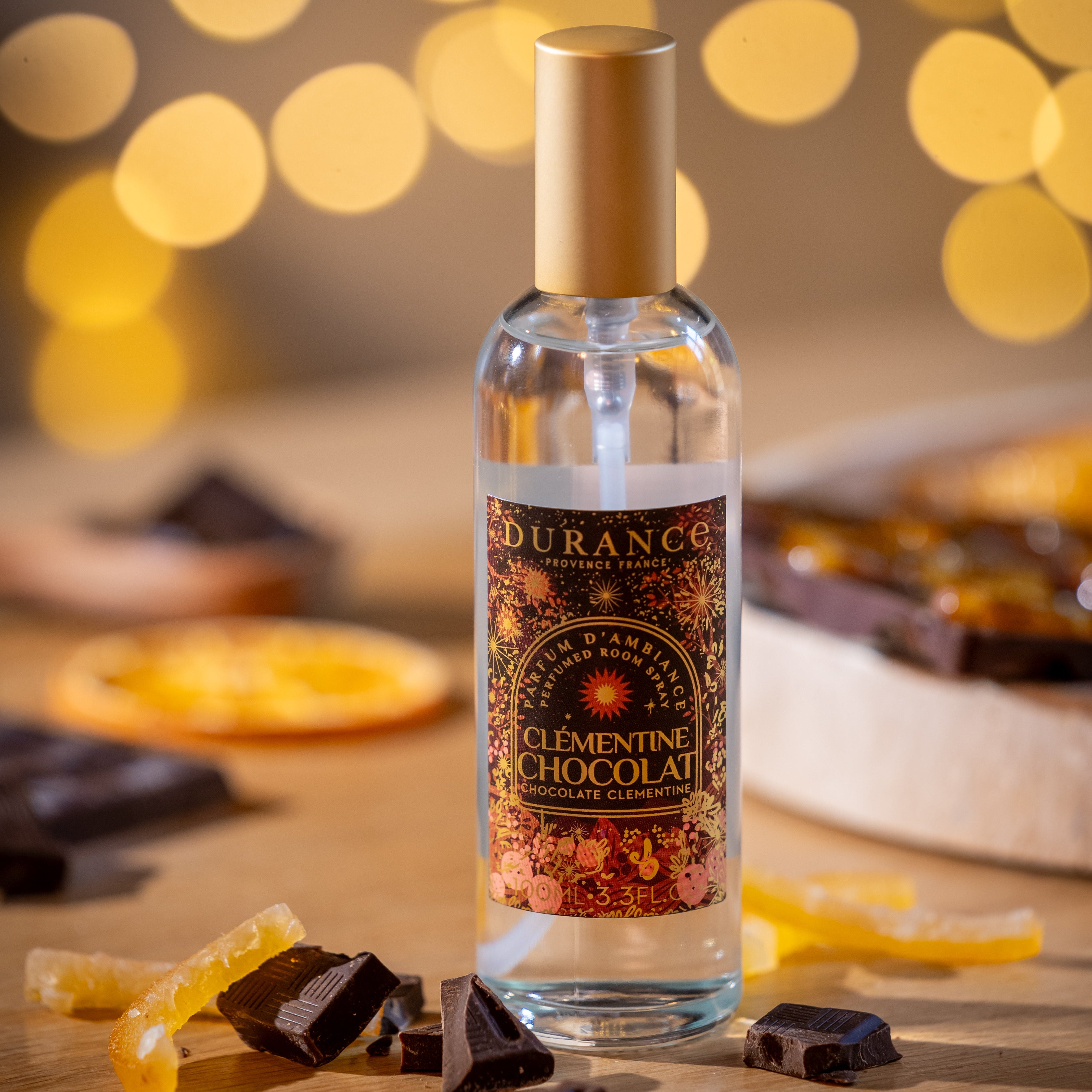 Bottle of Durance Clementine Chocolat on a wooden surface with chocolate and orange slices, blurred lights in the background