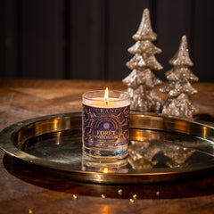 Candle in a decorative glass holder on a metallic tray with Christmas trees in the background.
