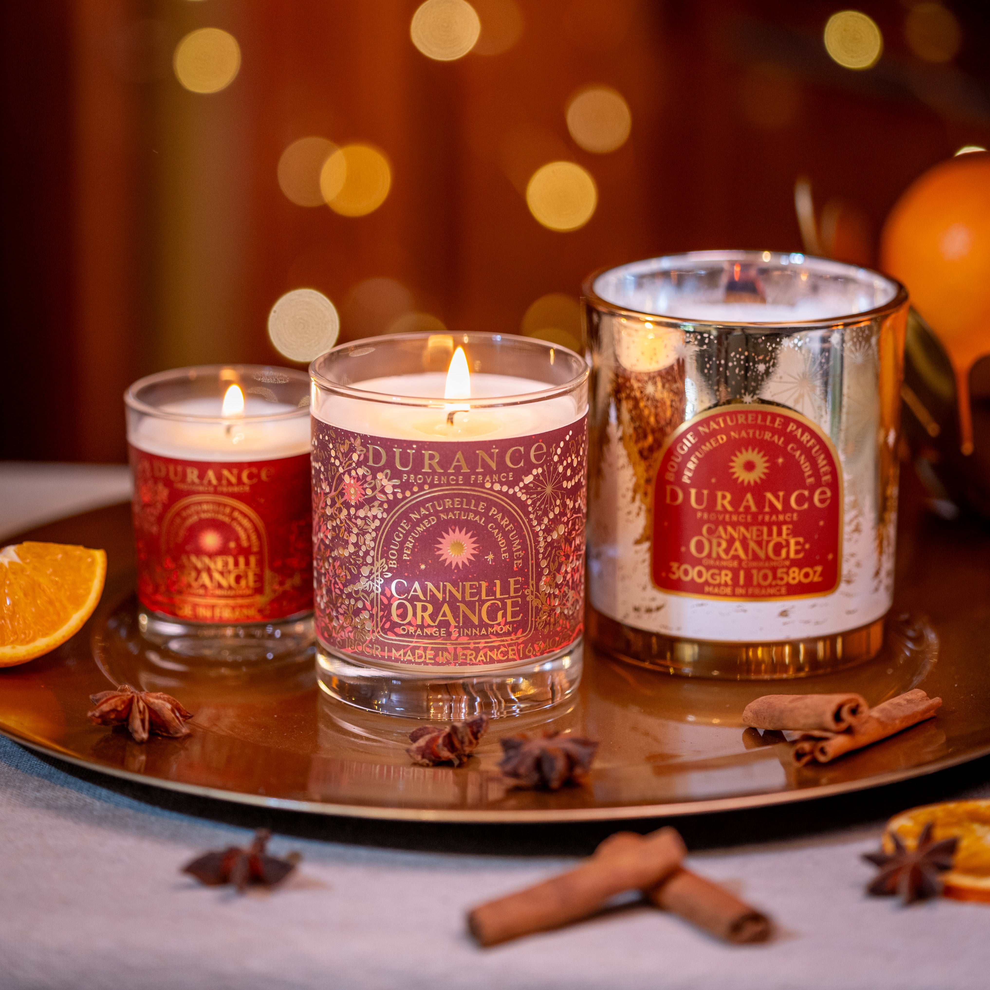 Three Durance candles on a tray with oranges and cinnamon sticks, blurred lights in the background