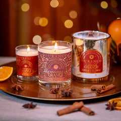 Three Durance candles on a tray with oranges and cinnamon sticks, blurred lights in the background