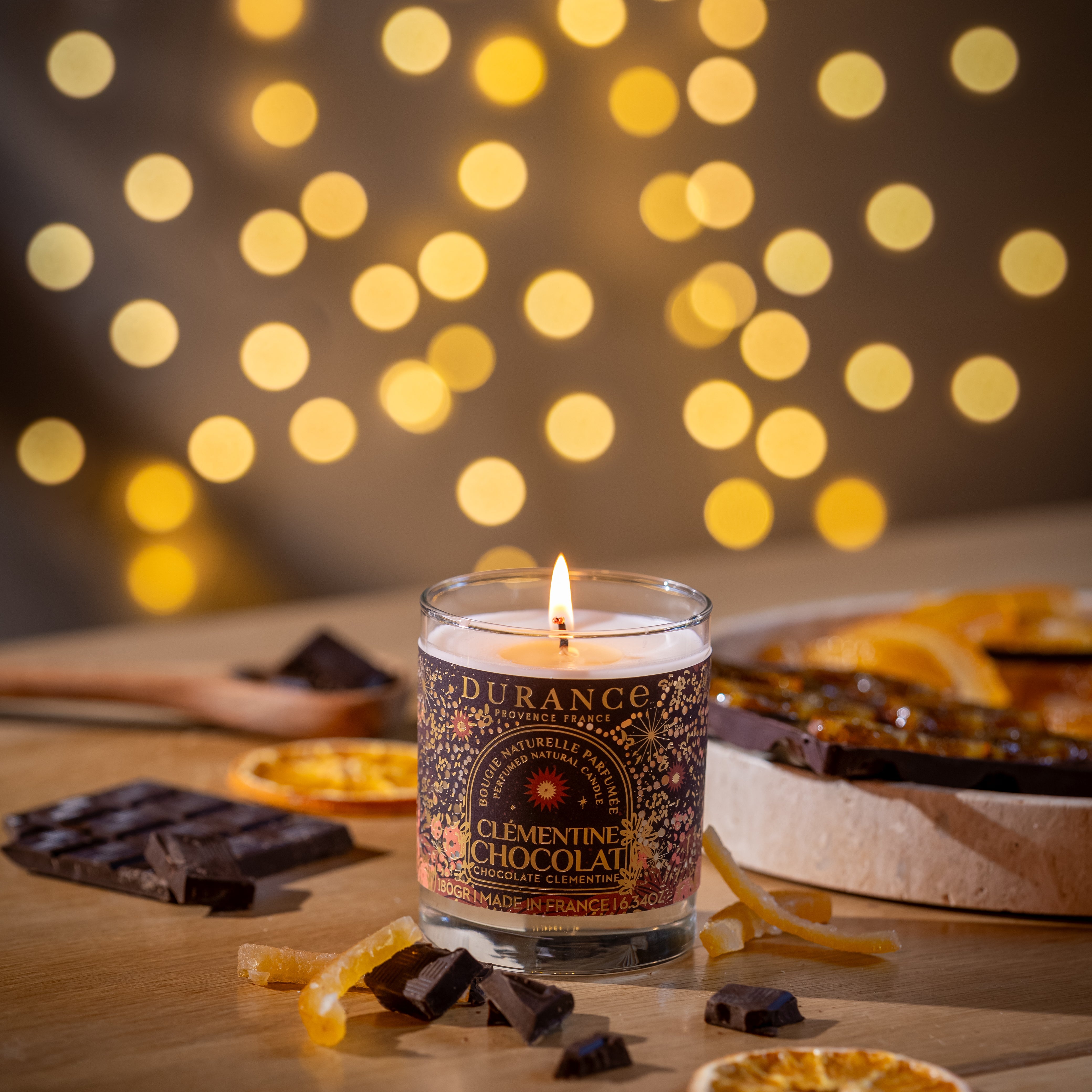 Candle with decorative packaging on a wooden surface with oranges and chocolate, blurred lights in the background