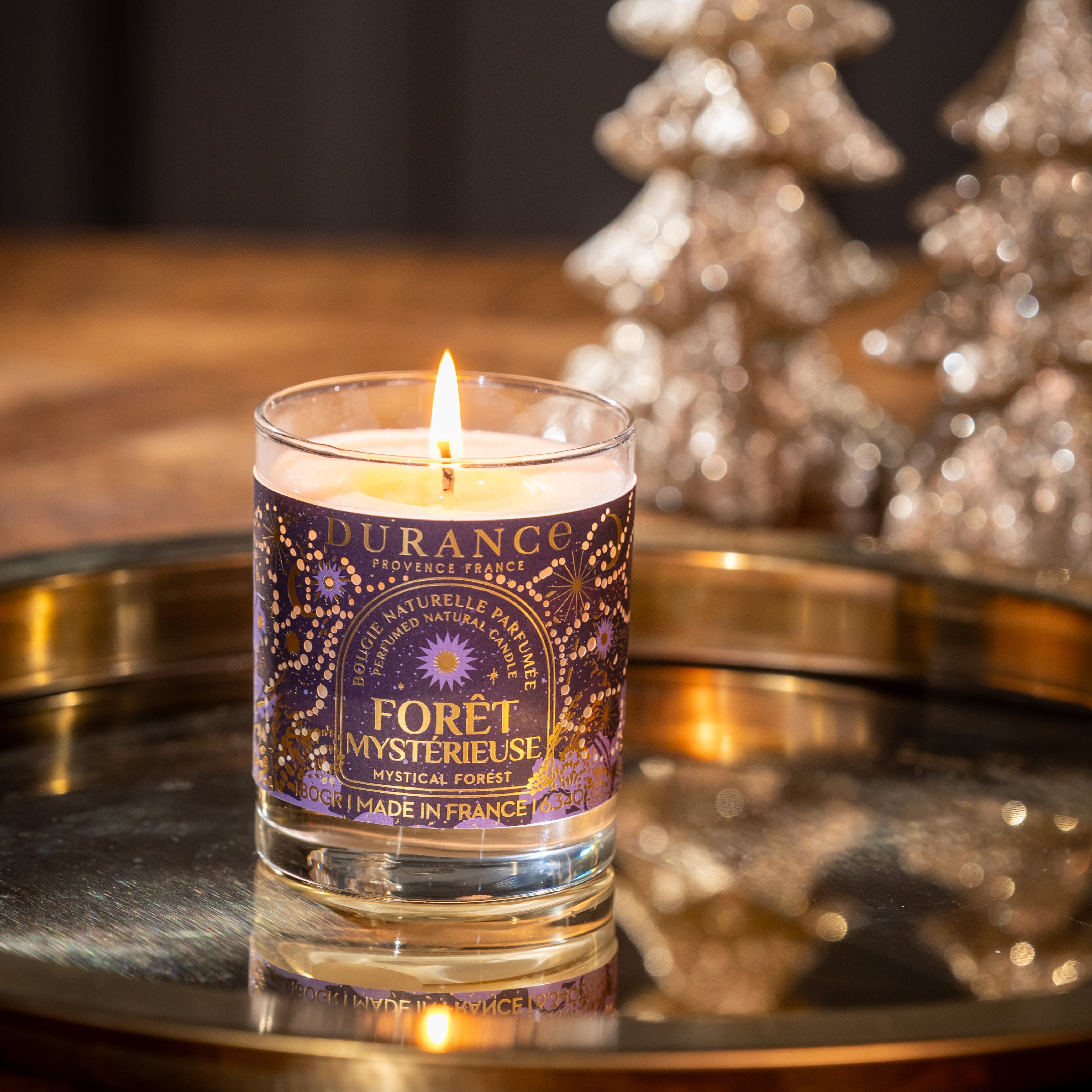 Candle in a glass holder with a purple label on a reflective surface, with decorative trees in the background.