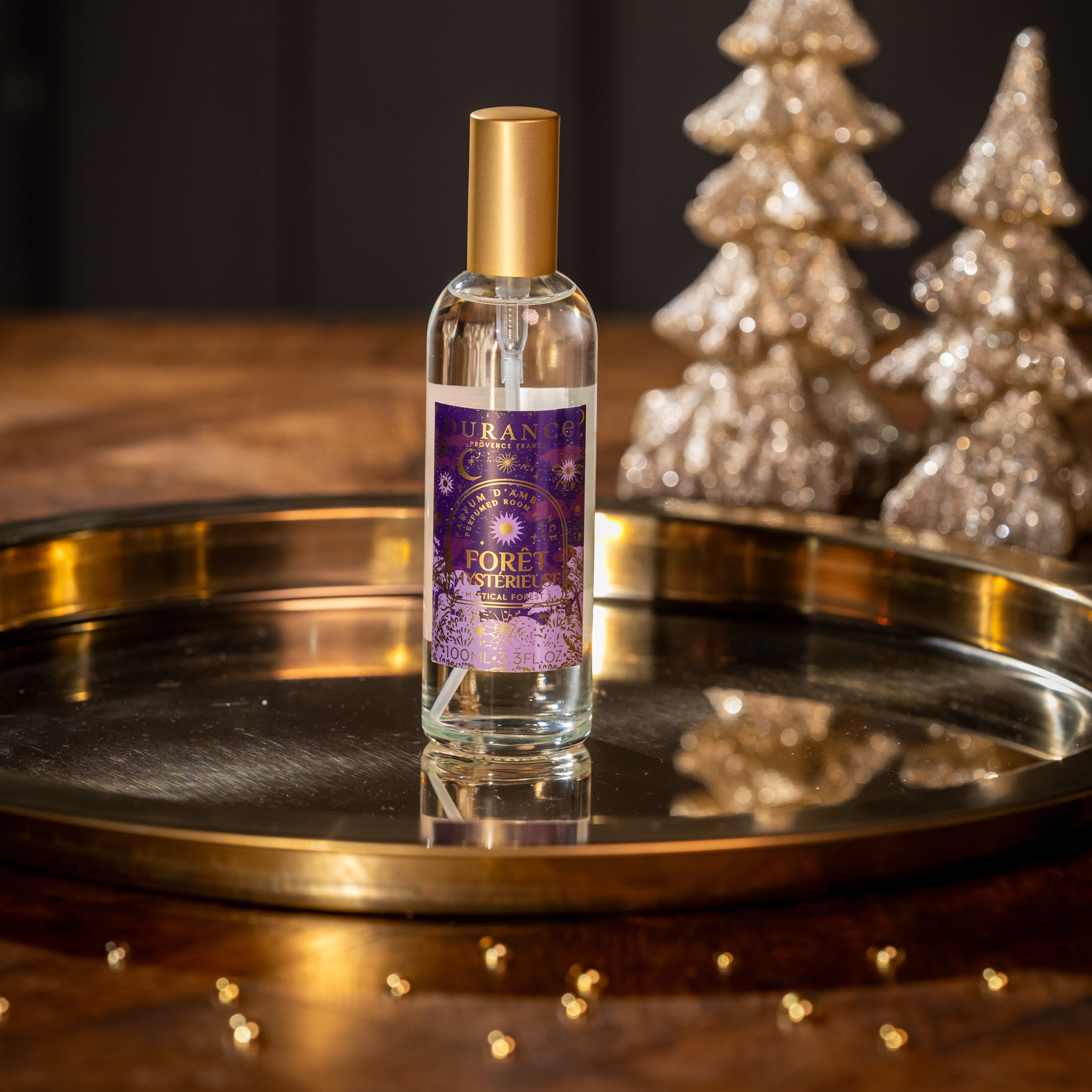 Bottle of perfume with a gold cap on a reflective surface with decorative trees in the background.