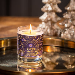 Candle in a glass holder with a purple label on a reflective surface, with decorative trees in the background.