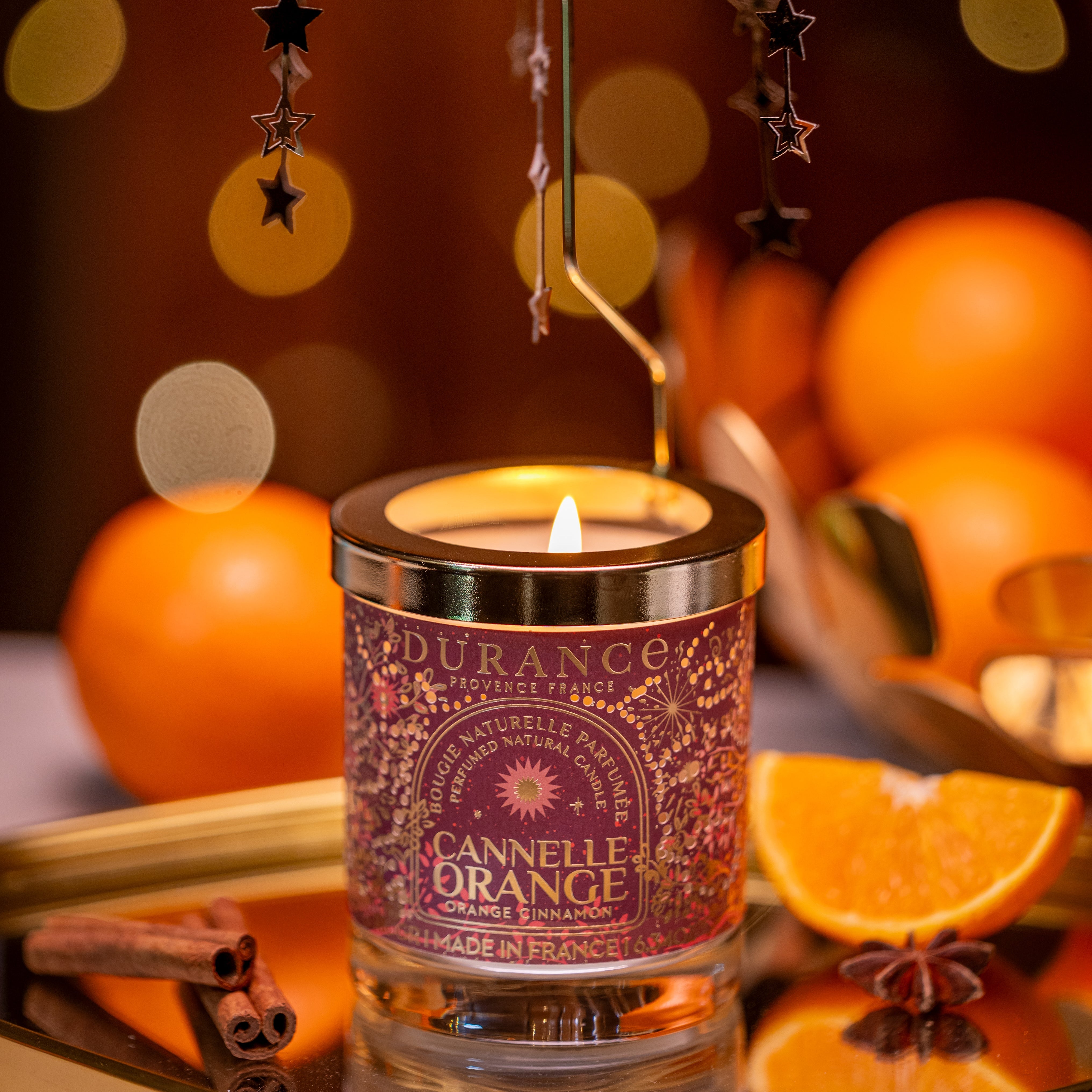 Candle labeled 'Cannelle Orange' with star-shaped earrings on a decorative tray with oranges and cinnamon sticks.