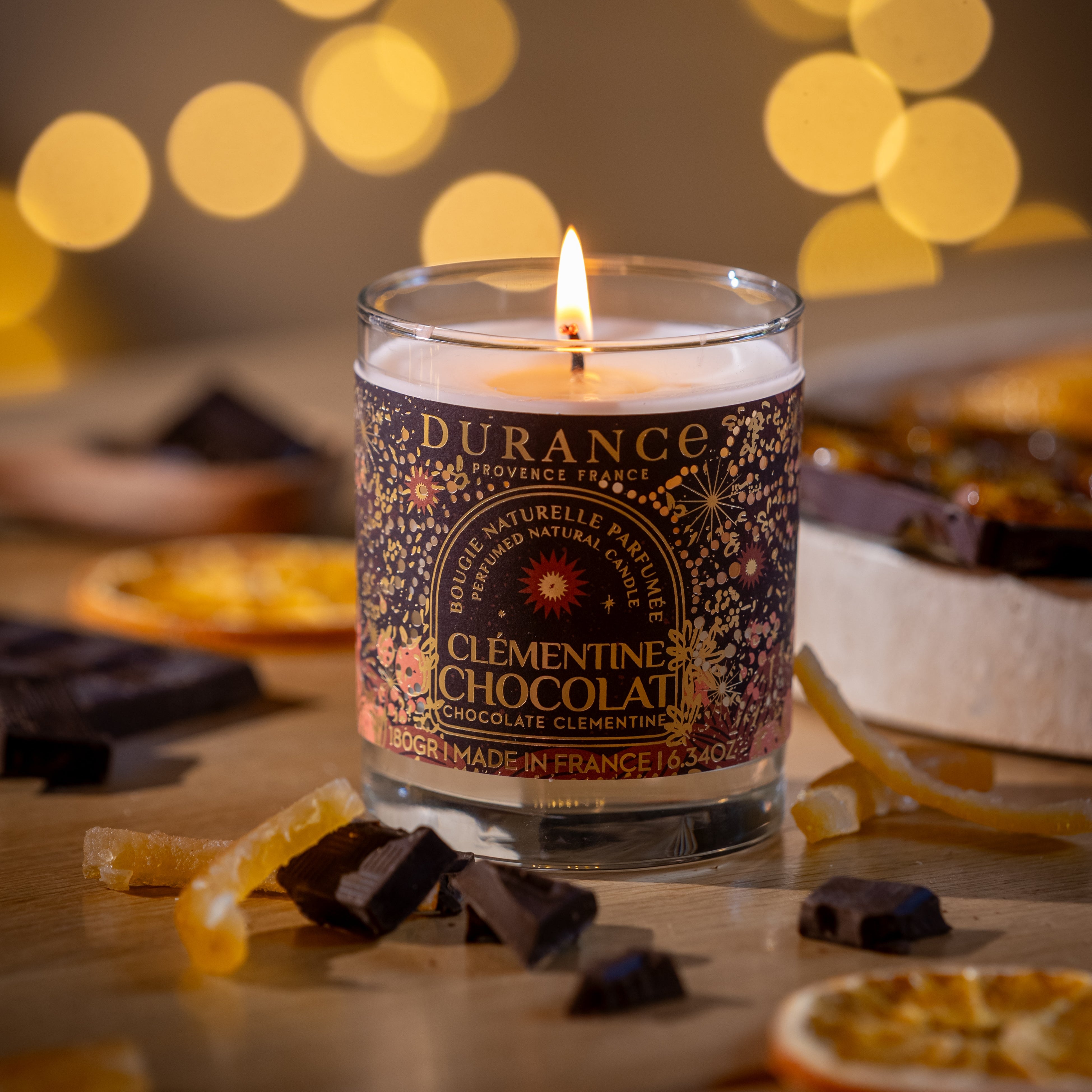 Durance Clementine Chocolatier candle with decorative packaging on a wooden surface with chocolate and orange slices, blurred lights in the background.