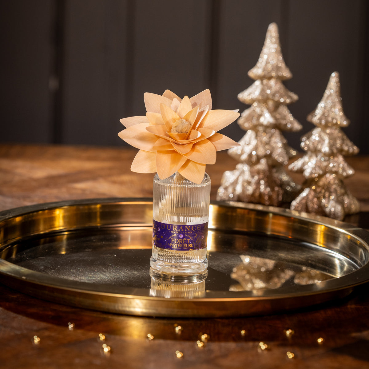 Decorative flower in a glass on a gold tray with metallic Christmas trees in the background.